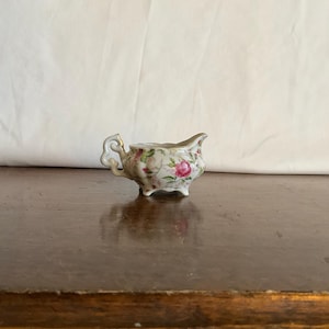 May include: A small, antique porcelain creamer with a floral rose pattern. The creamer has a delicate handle and spout, and rests on three small feet. The design features pink roses and green leaves on a white background, with gold trim.