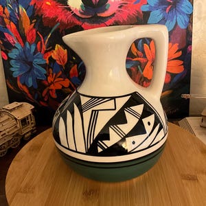 May include: A white ceramic pitcher with a black and white geometric design on the lower half. The base is green, and the pitcher has a curved handle. The background features a colorful painting of a panda.