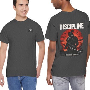 Può includere: T-shirt grigio scuro con un simbolo giapponese sul davanti e una grafica di un guerriero samurai sul retro. La grafica sul retro presenta una silhouette di samurai contro un tramonto rosso con la parola "DISCIPLINE" sopra e "BUSHIDO CODE" sotto.