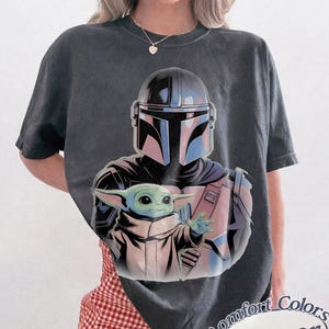 May include: Dark gray t-shirt featuring a graphic of The Mandalorian and Grogu. The Mandalorian is in a helmet and armor, holding Grogu. The shirt has the words "Comfort Colors Pepper" in a circular design.
