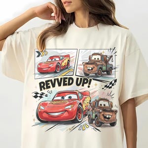May include: An ivory t-shirt featuring a graphic print of Lightning McQueen and Mater from the movie Cars. The design includes two smaller panels at the top and a larger central illustration with the text REVVED UP! and checkered flag accents.