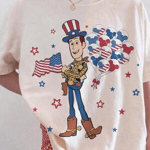 May include: Ivory-coloured t-shirt featuring a cartoon cowboy holding an American flag and balloons. The design includes red, white, and blue stars and balloons. The shirt has the words "Comfort Colors Ivory" printed on it.