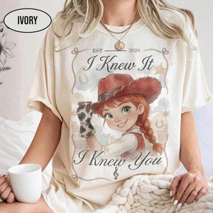 May include: An ivory short-sleeve t-shirt featuring a graphic of a cartoon cowgirl with red hair and a brown hat. The text reads "EST 2026", "I Knew It", and "I Knew You" with a musical note symbol. The design includes a rope border and stars.