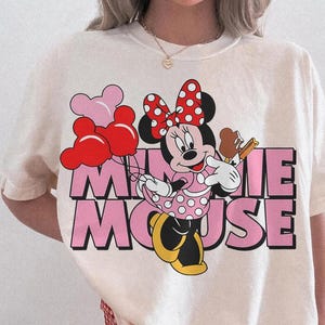 May include: Ivory-colored t-shirt featuring a graphic of Minnie Mouse holding balloons and an ice cream cone. The text "MINNIE MOUSE" is in pink block letters. The shirt is labeled "Comfort Colors Ivory".