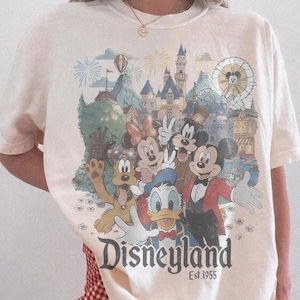 Puede incluir: Camiseta color marfil con un dise&ntilde;o de Disneyland que presenta a Mickey Mouse, Minnie Mouse, Donald Duck y Goofy. El dise&ntilde;o incluye un castillo, fuegos artificiales y una noria. El texto "Disneyland Est. 1955" est&aacute; impreso debajo.