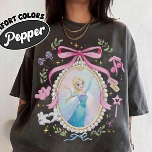 May include: A charcoal gray t-shirt featuring a graphic print of a blonde character in a blue dress inside a pearl-bordered oval frame. The design includes a pink bow, white cartoon gloves forming a heart, and small star and cherry accents.