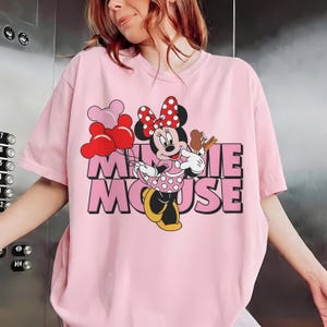 May include: Pink t-shirt featuring a Minnie Mouse graphic. The design includes Minnie holding balloons and a treat, with the words "Minnie Mouse" in pink and black block letters. The shirt has short sleeves.