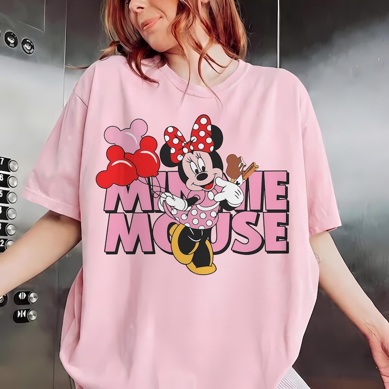 May include: Pink t-shirt featuring a Minnie Mouse graphic. The design includes Minnie holding balloons and a treat, with the words "Minnie Mouse" in pink and black block letters. The shirt has short sleeves.