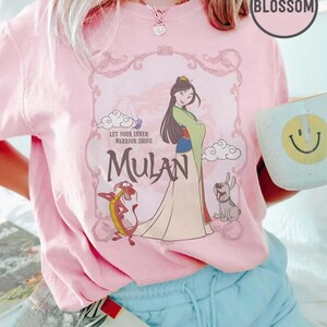 May include: Pink t-shirt featuring a graphic of Mulan with Mushu and a dog, with the text "Let Your Inner Warrior Shine" and "Mulan". The shirt has a crew neck and short sleeves.