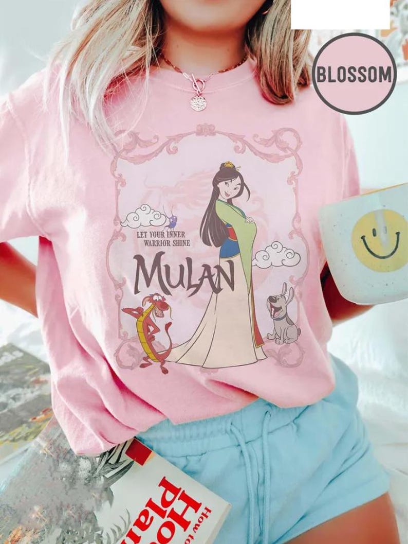 May include: Pink t-shirt featuring a graphic of Mulan with Mushu and a dog, with the text "Let Your Inner Warrior Shine" and "Mulan". The shirt has a crew neck and short sleeves.