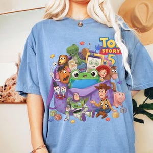 May include: A blue t-shirt featuring a graphic print of Toy Story 5 characters including Woody, Buzz Lightyear, Jessie, Rex, Slinky Dog, Mr. Potato Head, Forky, and a green frog emerging from a purple backpack, surrounded by colorful stars and blocks.