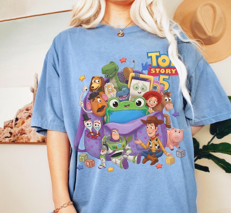 May include: A blue t-shirt featuring a graphic print of Toy Story 5 characters including Woody, Buzz Lightyear, Jessie, Rex, Slinky Dog, Mr. Potato Head, Forky, and a green frog emerging from a purple backpack, surrounded by colorful stars and blocks.