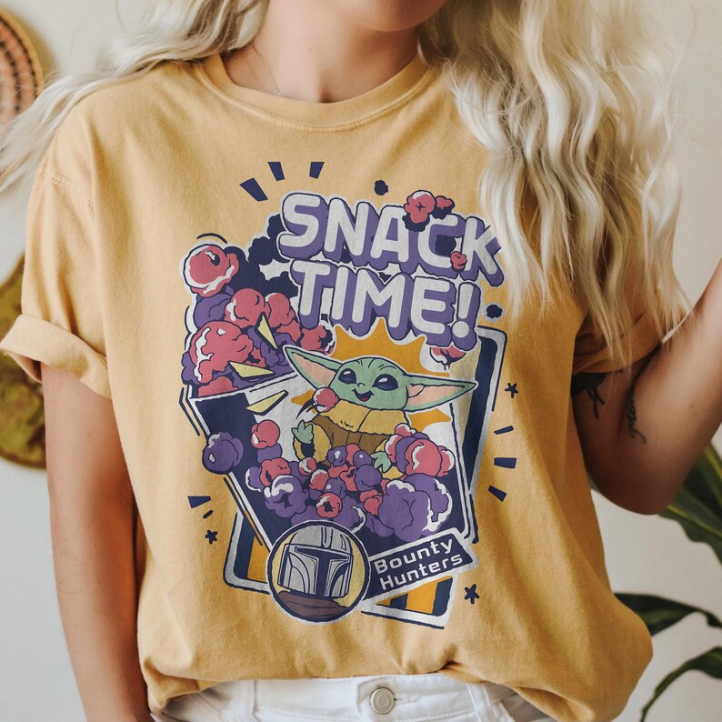 May include: A mustard yellow t-shirt featuring a graphic print with the text "SNACK TIME!" and "BOUNTY HUNTERS". The design depicts a green alien character surrounded by purple and pink clouds, with a helmet icon in a circular frame below.