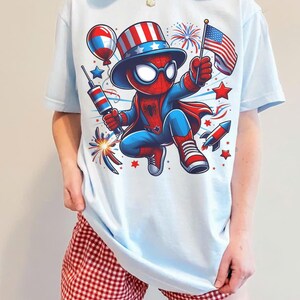 K&ouml;nnte beinhalten: Hellblaues T-Shirt mit einer Cartoon-Spider-Man-Figur in einem patriotischen Outfit, die ein Feuerwerk und eine amerikanische Flagge h&auml;lt. Das Design umfasst Feuerwerk, Sterne und eine Rakete. Die Person tr&auml;gt rot-wei&szlig; karierte Shorts.