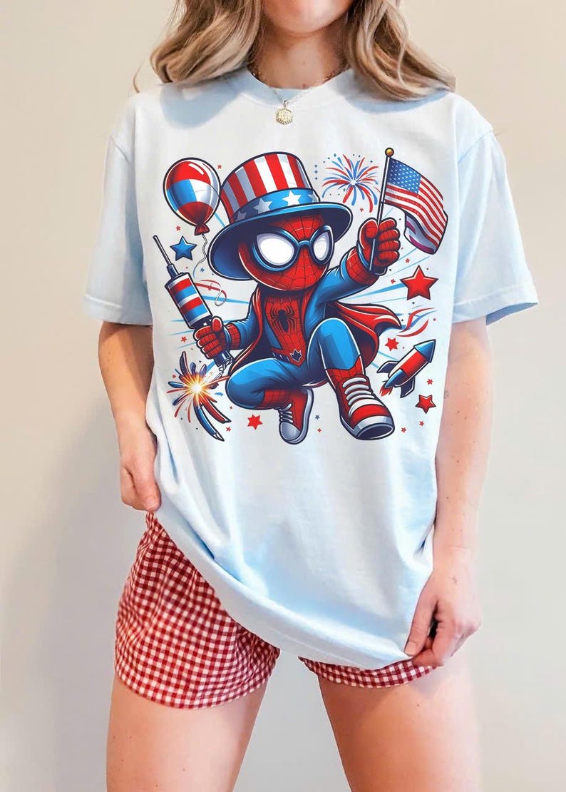 K&ouml;nnte beinhalten: Hellblaues T-Shirt mit einer Cartoon-Spider-Man-Figur in einem patriotischen Outfit, die ein Feuerwerk und eine amerikanische Flagge h&auml;lt. Das Design umfasst Feuerwerk, Sterne und eine Rakete. Die Person tr&auml;gt rot-wei&szlig; karierte Shorts.