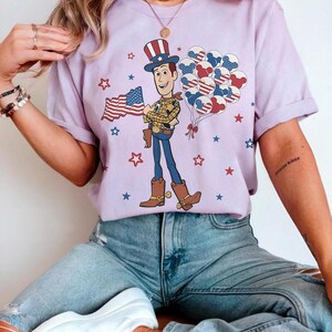May include: A lilac t-shirt with a cartoon cowboy holding a US flag and balloons. The cowboy is wearing a hat, vest, and boots. Red, white, and blue stars and balloons decorate the shirt, creating a patriotic theme.