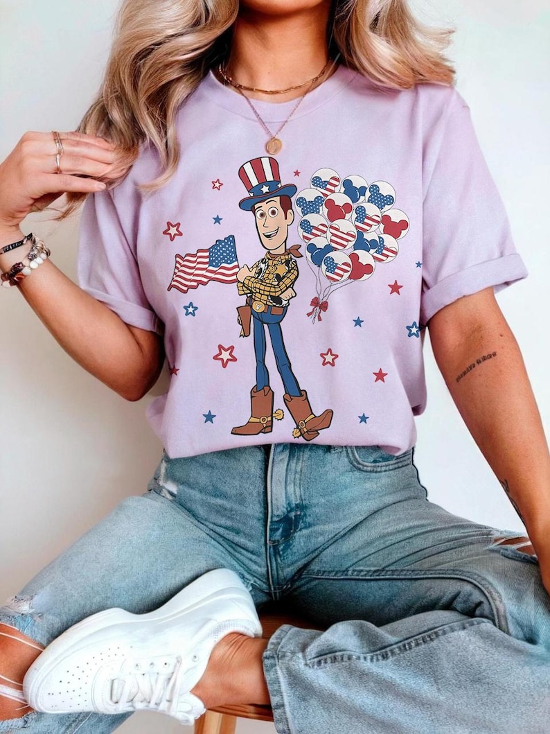 May include: A lilac t-shirt with a cartoon cowboy holding a US flag and balloons. The cowboy is wearing a hat, vest, and boots. Red, white, and blue stars and balloons decorate the shirt, creating a patriotic theme.