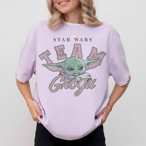 Puede incluir: Una camiseta de manga corta de color morado claro con un estampado gr&aacute;fico del personaje Grogu. El texto STAR WARS aparece sobre el personaje y TEAM GROGU en letras grandes de estilo desgastado alrededor de la ilustraci&oacute;n verde.