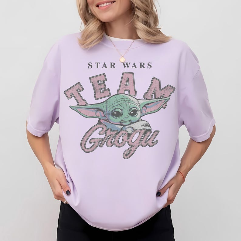Puede incluir: Una camiseta de manga corta de color morado claro con un estampado gr&aacute;fico del personaje Grogu. El texto STAR WARS aparece sobre el personaje y TEAM GROGU en letras grandes de estilo desgastado alrededor de la ilustraci&oacute;n verde.