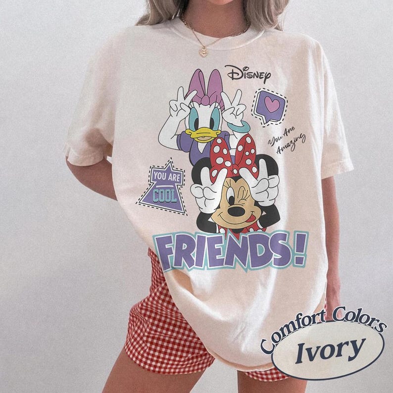 May include: Ivory-colored t-shirt featuring Disney's Minnie Mouse and Daisy Duck. The graphic includes the word "FRIENDS!" in purple, along with the phrases "You Are Cool" and "You Are Amazing." The shirt is labeled "Comfort Colors Ivory."