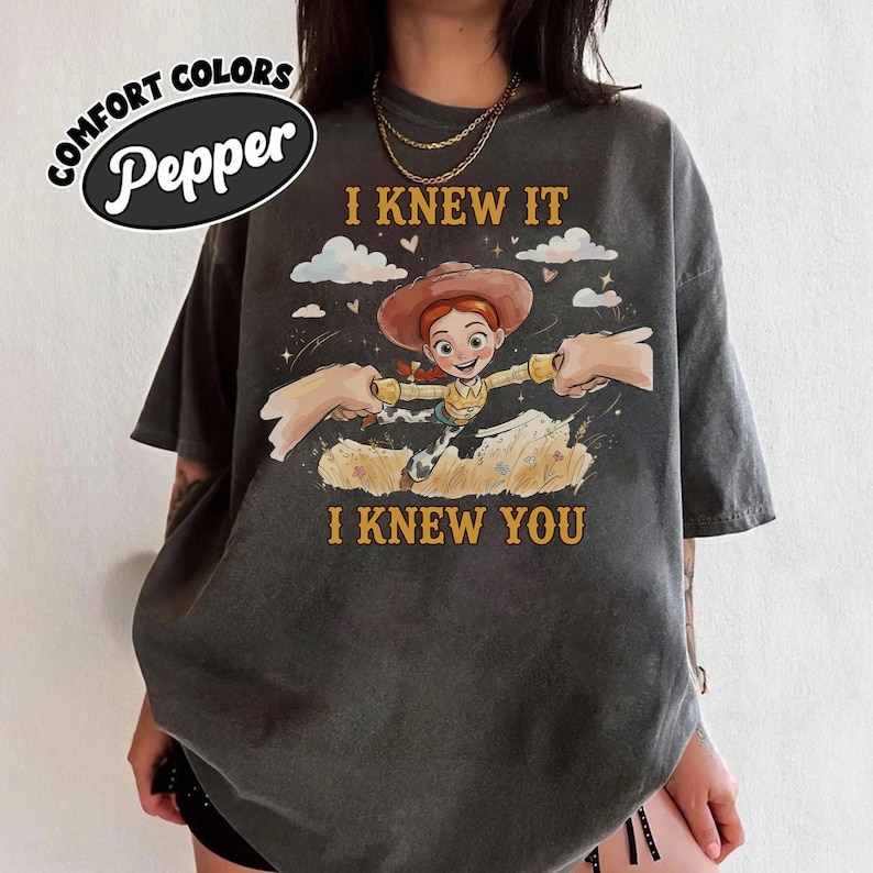 May include: A charcoal gray t-shirt featuring a graphic of a cartoon cowgirl character with red hair and a brown hat. The text reads I KNEW IT and I KNEW YOU. The design includes clouds, stars, and two hands reaching toward the character. The Comfort Colors Pepper brand label is visible in the upper left corner.