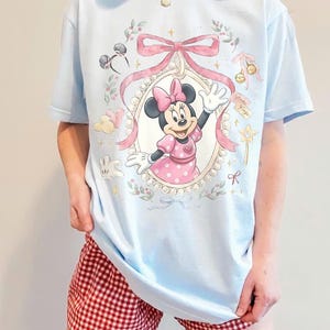 May include: A light blue t-shirt featuring a graphic of Minnie Mouse inside a pearl-bordered oval frame, accented with a large pink bow, floral sprigs, a star wand, and small icons including mouse ears and balloons, all printed in soft pastel colors.