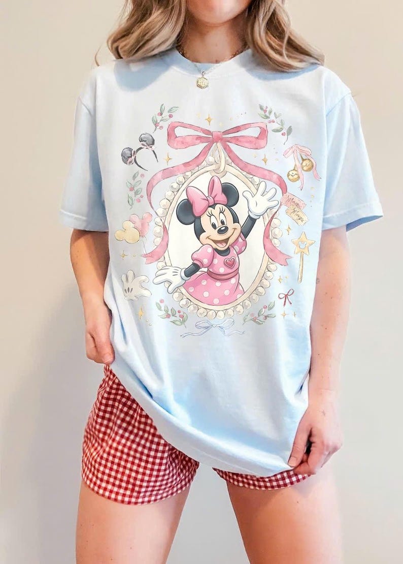 May include: A light blue t-shirt featuring a graphic of Minnie Mouse inside a pearl-bordered oval frame, accented with a large pink bow, floral sprigs, a star wand, and small icons including mouse ears and balloons, all printed in soft pastel colors.