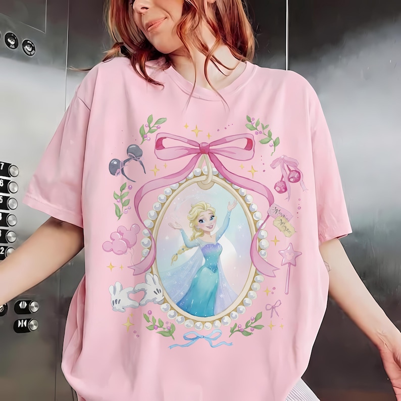 May include: A light pink t-shirt featuring a central oval frame decorated with white pearls and a large pink bow. Inside the frame is an illustration of a blonde character in a blue dress. Surrounding elements include mouse ears, balloons, and stars.
