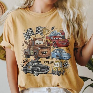 May include: A mustard yellow t-shirt featuring a graphic print of characters from the movie Cars, including a brown tow truck, a red race car with the number 95, a blue sports car, a black police car, and a yellow forklift, with the text GO and SPEED.
