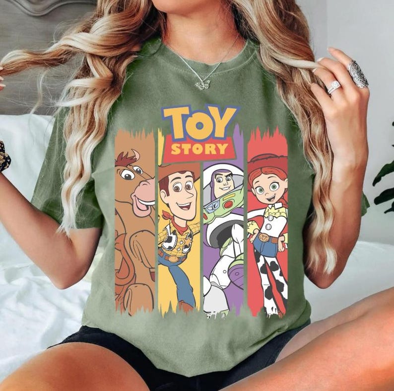 May include: Green t-shirt featuring the Toy Story logo and characters. The design includes images of Bullseye, Woody, Buzz Lightyear, and Jessie. The shirt has a classic crew neck and short sleeves.