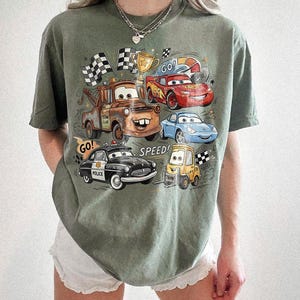 May include: An olive green t-shirt featuring a graphic print of characters from the movie Cars, including a brown tow truck, a red race car, a blue sports car, a black police car, and a yellow forklift, with the text GO and SPEED.