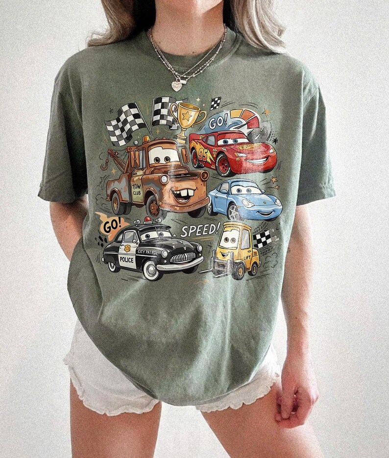 May include: An olive green t-shirt featuring a graphic print of characters from the movie Cars, including a brown tow truck, a red race car, a blue sports car, a black police car, and a yellow forklift, with the text GO and SPEED.