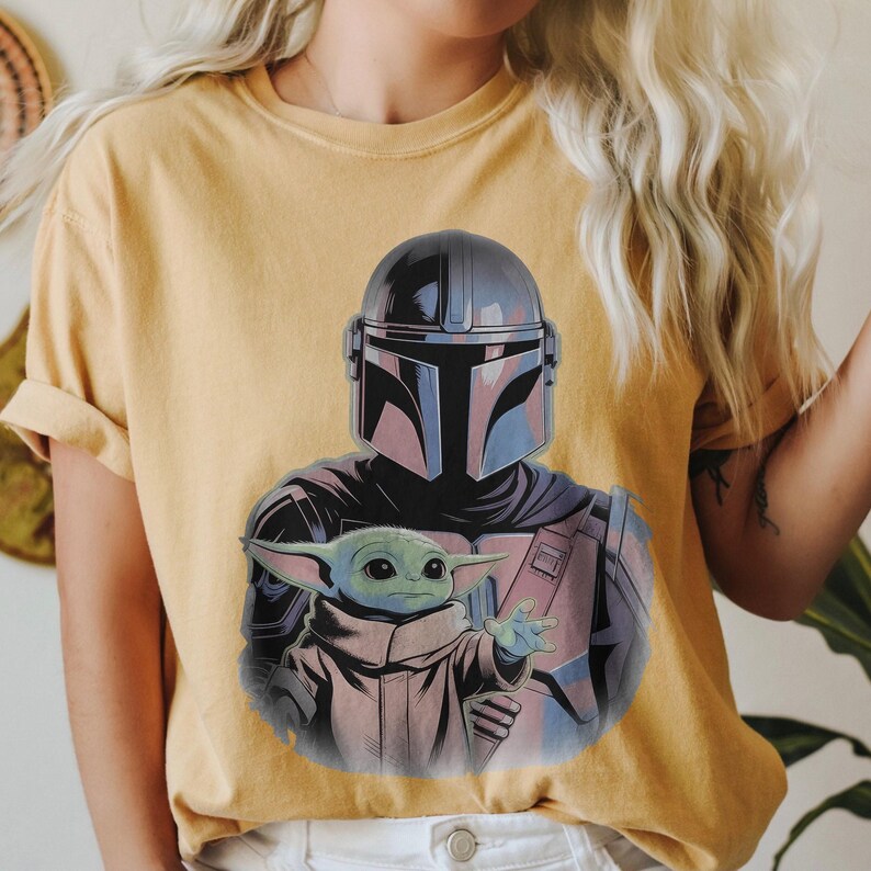 May include: Mustard yellow t-shirt featuring a graphic of The Mandalorian and Grogu. The Mandalorian's helmet is black and blue, and Grogu is green. The graphic is centered on the shirt.