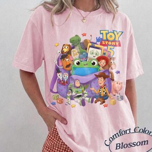 May include: A light pink t-shirt featuring a graphic print of various Toy Story characters including Woody, Buzz Lightyear, and a green frog emerging from a purple backpack, with the text Toy Story 5 printed in blue and yellow above the characters.