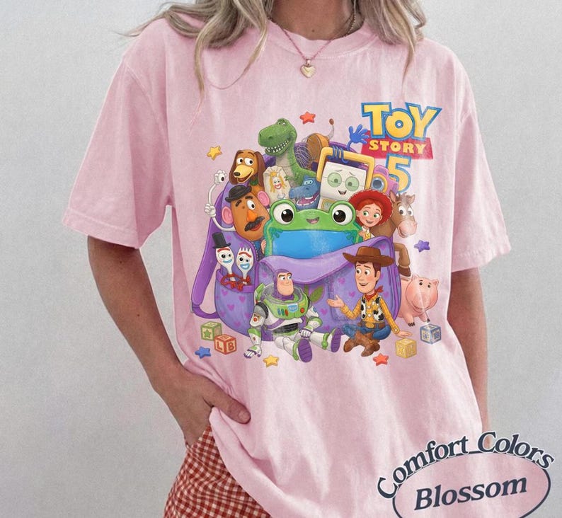 May include: A light pink t-shirt featuring a graphic print of various Toy Story characters including Woody, Buzz Lightyear, and a green frog emerging from a purple backpack, with the text Toy Story 5 printed in blue and yellow above the characters.
