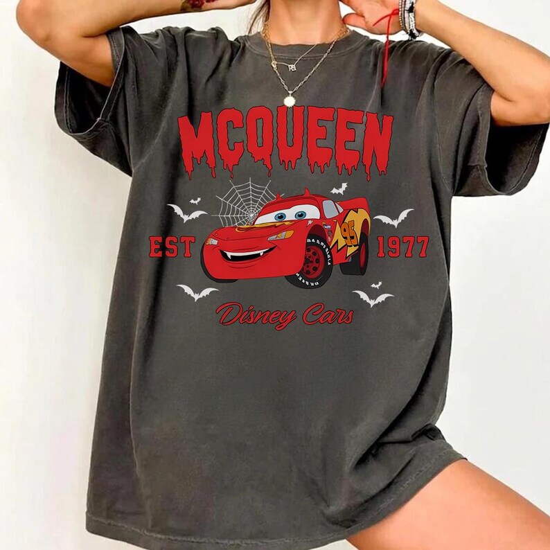 May include: Dark gray t-shirt featuring a red graphic of Lightning McQueen from Disney Cars. The graphic includes the text "McQueen", "EST 1977", and "Disney Cars", along with bats and a spiderweb.
