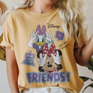 Może przedstawiać: Musztardowa koszulka z Daisy Duck i Minnie Mouse pokazującymi znak pokoju. Koszulka marki Disney zawiera napisy "You Are Cool", "You Are Amazing" i "FRIENDS!" w fioletowym i białym kolorze.