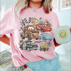 May include: A light pink t-shirt featuring a graphic print of various cartoon vehicles including a brown tow truck, a red race car, a blue sports car, a black police car, and a yellow forklift, with the text GO and SPEED and checkered flags.