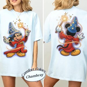 May include: Pale blue t-shirt with a front and back graphic of Mickey Mouse dressed as a sorcerer, holding a wand with stars. The shirt has short sleeves and a loose fit. The text "Comfort Colors Chambray" is on the bottom.