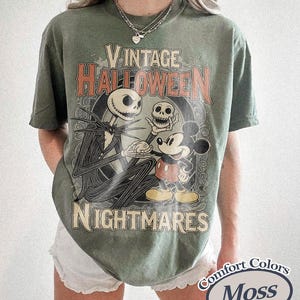 May include: A moss green short-sleeve t-shirt featuring a graphic print with the text VINTAGE HALLOWEEN NIGHTMARES. The design depicts a skeletal character holding a skull and standing next to a cartoon mouse character in a circular frame.