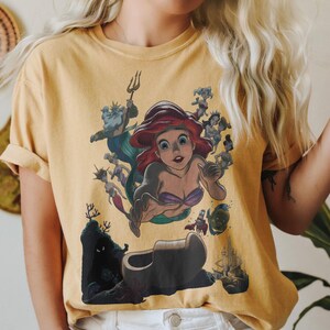 May include: A mustard yellow short-sleeve t-shirt featuring a graphic print of a red-haired mermaid character surrounded by various underwater figures, including a king with a trident, a crab, a fish, and a castle silhouette at the bottom.