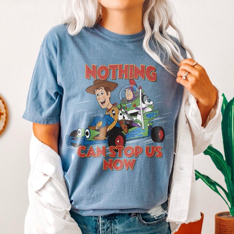 以下が含まれることがあります： トイ・ストーリーのウッディとバズ・ライトイヤーがカートに乗った青いTシャツ。「NOTHING CAN STOP US NOW」の文字が赤色でキャラクターの上と下にプリントされています。Tシャツは半袖です。