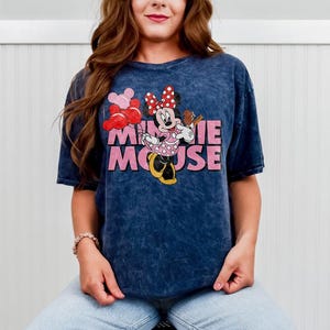 May include: A navy blue t-shirt featuring a graphic of Minnie Mouse holding balloons and a treat. The words "Minnie Mouse" are printed in pink. The shirt has a vintage, washed appearance.