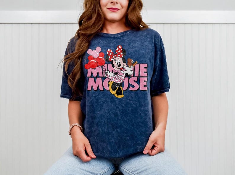 May include: A navy blue t-shirt featuring a graphic of Minnie Mouse holding balloons and a treat. The words "Minnie Mouse" are printed in pink. The shirt has a vintage, washed appearance.