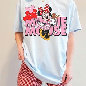 May include: Light blue t-shirt featuring a Minnie Mouse graphic. The graphic includes Minnie Mouse holding a treat and balloons, with the words "MINNIE MOUSE" in pink. The person is also wearing red and white checkered shorts.