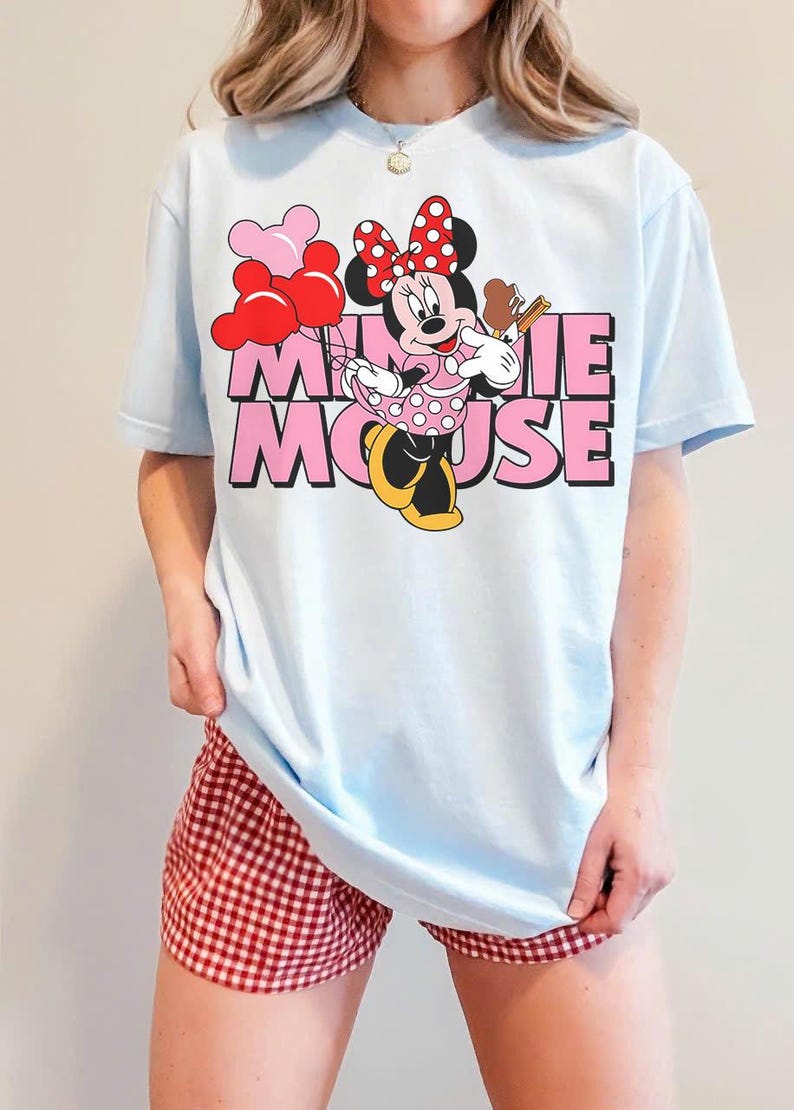 May include: Light blue t-shirt featuring a Minnie Mouse graphic. The graphic includes Minnie Mouse holding a treat and balloons, with the words "MINNIE MOUSE" in pink. The person is also wearing red and white checkered shorts.