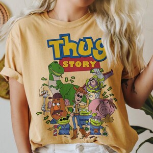 Pode incluir: Camiseta amarelo mostarda com um gr&aacute;fico de personagens de desenhos animados e o texto "Thug Story" em azul e amarelo. O gr&aacute;fico inclui um dinossauro verde, uma cabe&ccedil;a de batata e outros personagens.