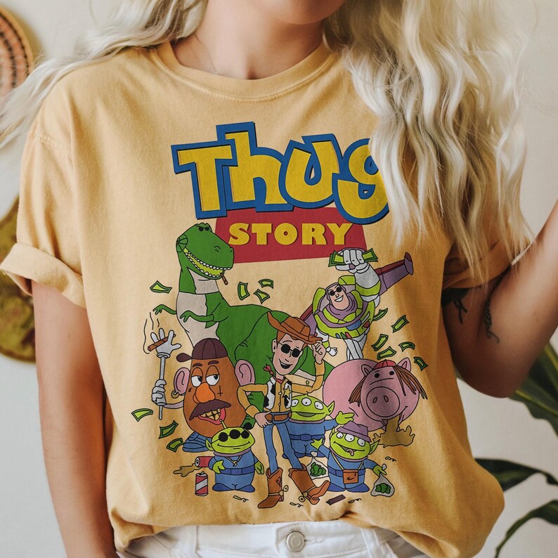Pode incluir: Camiseta amarelo mostarda com um gr&aacute;fico de personagens de desenhos animados e o texto "Thug Story" em azul e amarelo. O gr&aacute;fico inclui um dinossauro verde, uma cabe&ccedil;a de batata e outros personagens.