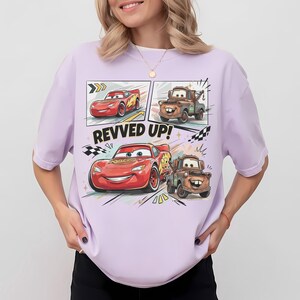 May include: A light purple t-shirt featuring a graphic print of the characters Lightning McQueen and Mater from the movie Cars. The design includes the text "REVVED UP!" along with checkered flag motifs and dynamic motion lines in a comic book style.
