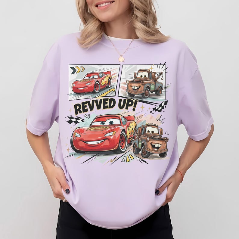 May include: A light purple t-shirt featuring a graphic print of the characters Lightning McQueen and Mater from the movie Cars. The design includes the text "REVVED UP!" along with checkered flag motifs and dynamic motion lines in a comic book style.